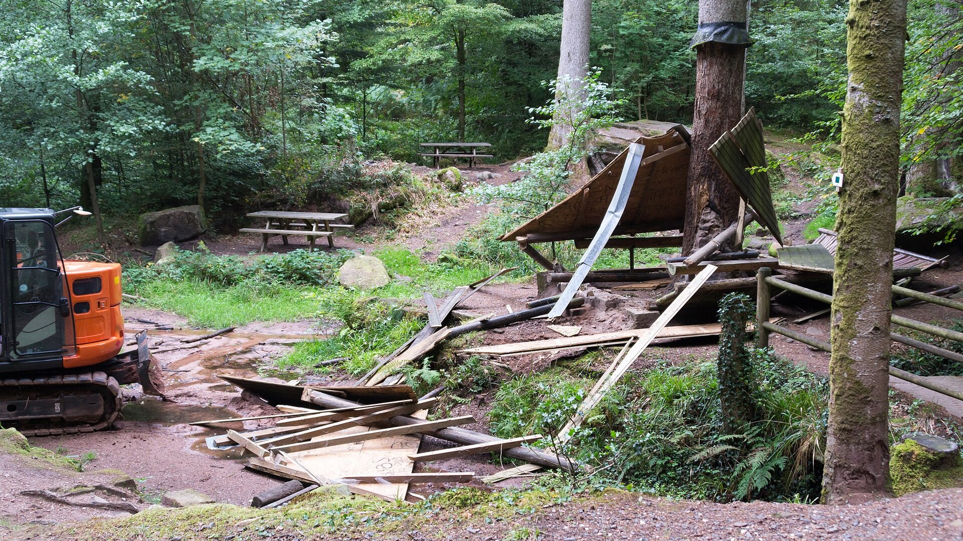 A small timber shelter flattened in woodland beside a parked orange digger (illustrative stock photo).