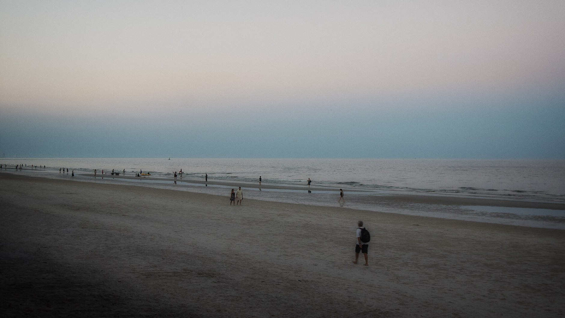 A few walkers on a wide, dim stretch of Hua Hin public beach at dusk, with a pale sky over the sea.