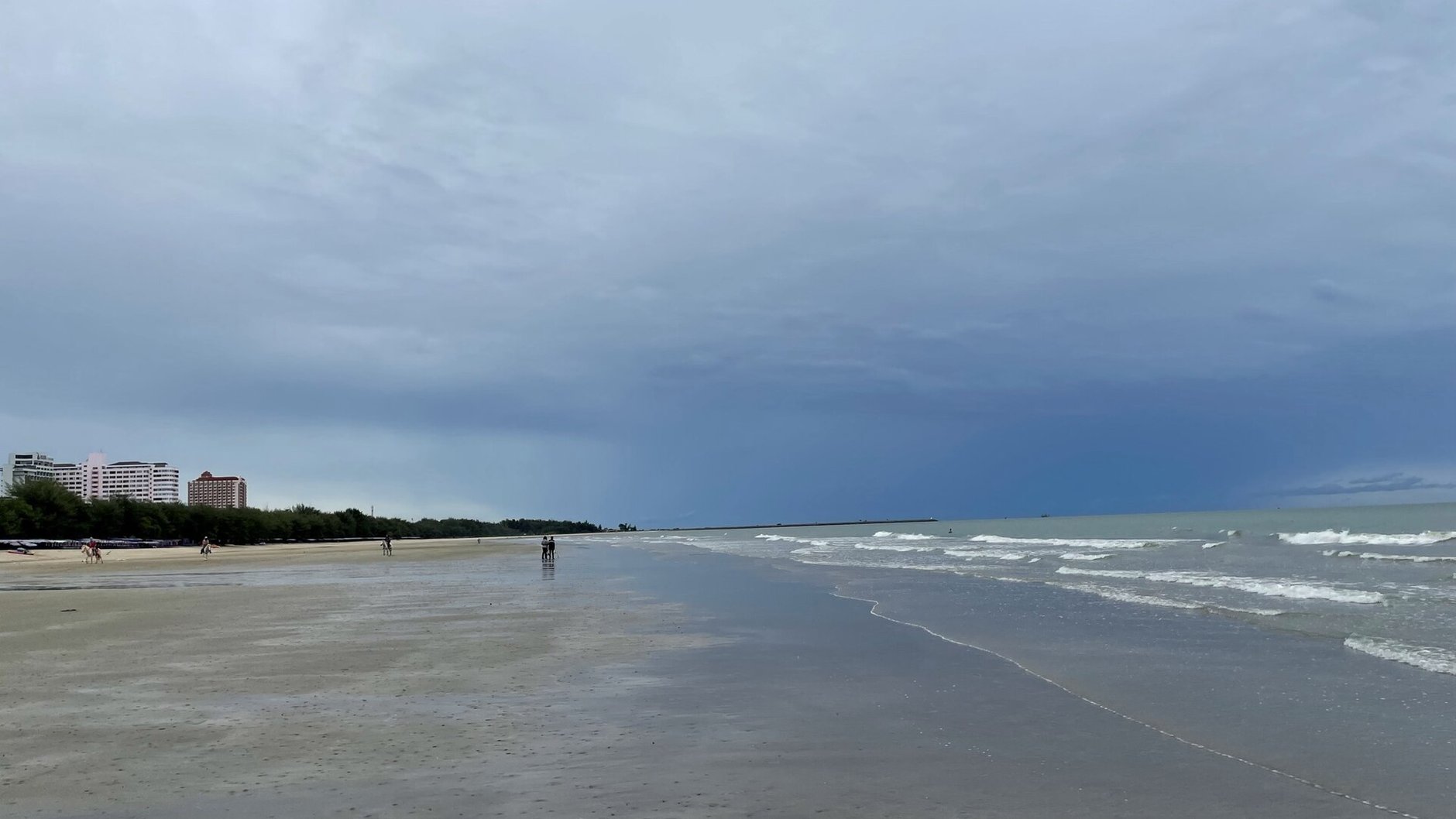 A wide, empty stretch of wet sand under a heavy grey sky, with small waves on the right and a few distant walkers.