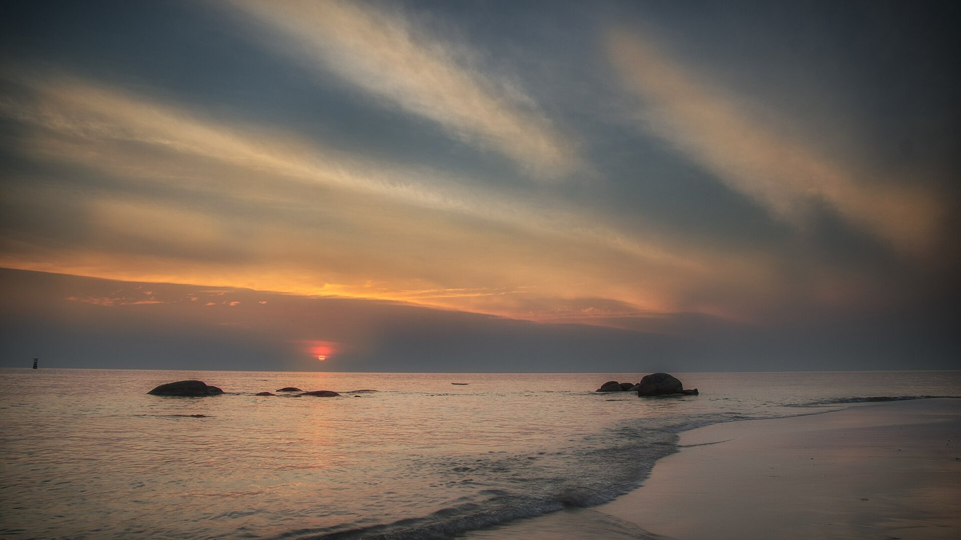 Low sun on the horizon over a calm sea at Hua Hin public beach, with rocks in the shallows and long streaks of cloud above.