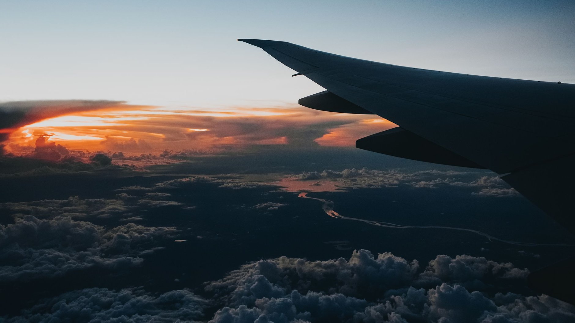 An aircraft wing silhouetted against an orange sunset above a layer of clouds (illustrative stock photo).