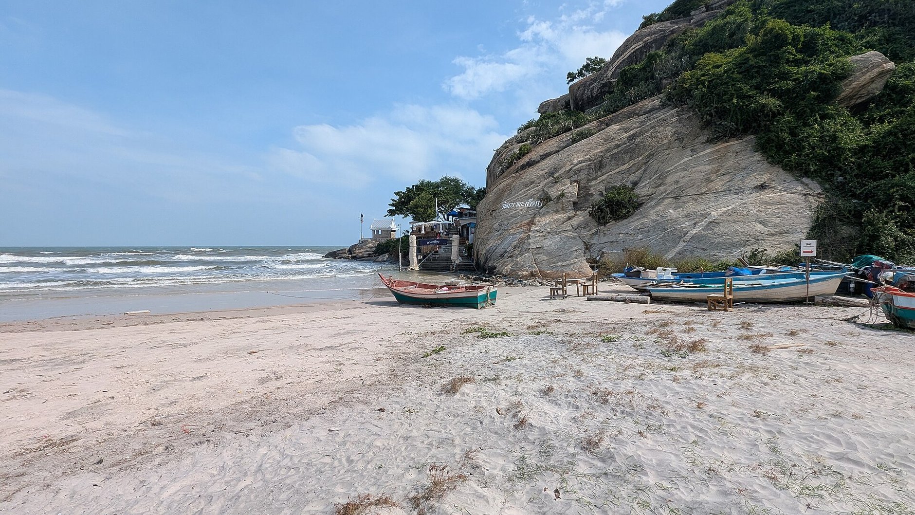 Painted fishing boats on pale sand below a granite headland, with small waves and blue sky beyond.